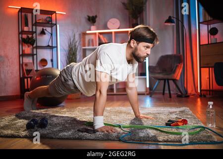 Endurance home workout. Young handsome strong active bearded man in sportswear doing push-ups exercises or standing on plank position on the floor at home in evening time Stock Photo