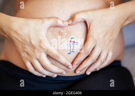 Painted boy on his belly of a pregnant woman. She creates a heart around the drawing with her hands. Stock Photo