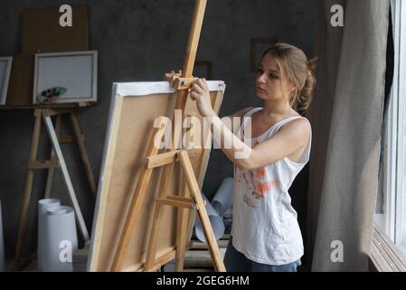 Portrait of a woman artist in an art studio stands near the easel and fixes the canvas. Stock Photo