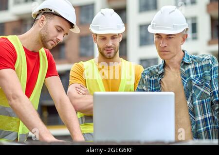 Three concentrated men in protective helmets standing before a laptop Stock Photo