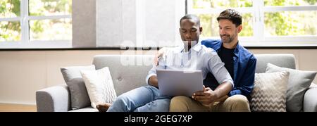 Gay Couple Using Computer For Video Conference On Sofa Or Couch Stock Photo