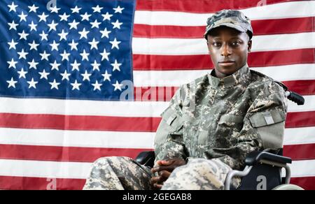 Patriotic Soldier Sitting In Wheel Chair Against American Flag Stock Photo