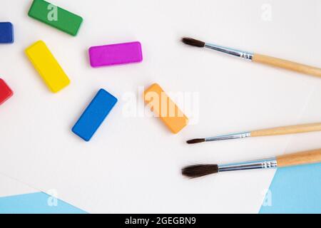 Multi-colored paints, watercolor brushes of different sizes lie together with watercolor paper on a blue background. Drawing lessons. View from above Stock Photo