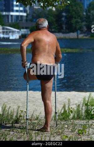 Old disabled one-legged man standing with double adjustable elbow crutches sunbathing on a river sandy beach. Stock Photo