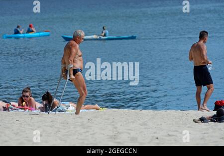 Old disabled one-legged man standing with double adjustable elbow crutches, people sunbathing on a river sandy beach, canoes floating. August 2, 2012. Stock Photo