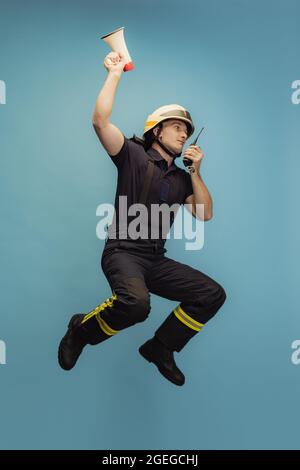 One young male fireman in uniform jumping with loudspeaker and walkie-talkie over blue background Stock Photo