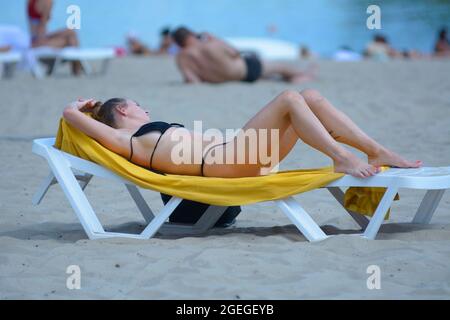 Young woman lying on the beach chaise lounge sunbathing on a sandy river beach, blurred people on a background. August 2, 2012. Kyiv, Ukraine Stock Photo