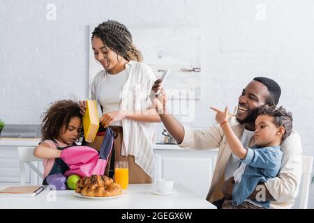 african american woman helping daughter packing backpack in kitchen Stock Photo