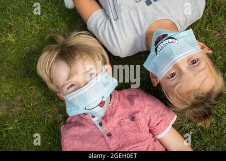 Portrait of two boys 5-6 years old in medical protective masks with funny smiles drawn. lie on backs on lawn, looking up. communication between childr Stock Photo