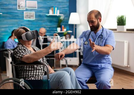 Male nurse giving social support, helping retired disabled senior woman to enjoy virtual reality using headset during caregiver service. Social nursing services at home. Healthcare assistance Stock Photo