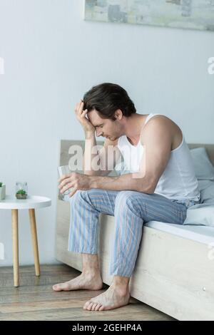 man in pajamas suffering from headache while sitting on bed with glass of water Stock Photo