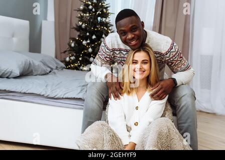 Lovely multiethnic couple are hugging at home against the background of the Christmas tree. African man hugs young Caucasian woman sitting on the floo Stock Photo