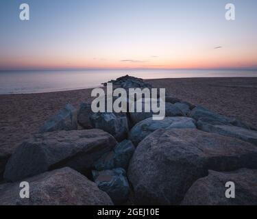 The view from a rock jetty on a sandy beach as the sun is rising over the Atlantic Ocean on Plum Island, Newburyport Massachusetts. The sky is pastel. Stock Photo