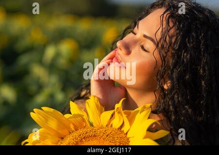 Close-up portrait of a young woman looking out from behind a large sunflower. Summertime concept. Stock Photo
