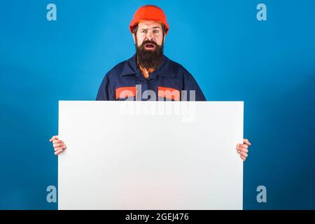 Confused builder with empty advertising banner. Bearded worker in protective clothing shows blank sign board ready for your text. Stock Photo