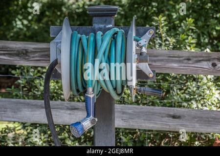 Heavy duty gardening hose with a hand sprayer attached coiled up on a metal reel that is mounted on a post against the fence in a garden for water sup Stock Photo