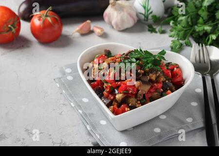 Salad with eggplants, tomatoes, garlic and parsley in a plate in the shape of a heart on a light gray background. Stock Photo