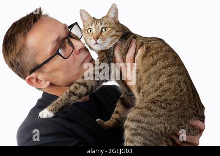 A man in glasses holds a cat in his hands and looks at her attentively. The cat looks away warily. Isolated on white. Stock Photo