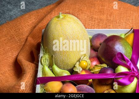 Juicy ripe fruits and berries are beautifully stacked in wooden box tied with pink ribbon. Dark background. Food delivery, gift fruit set. Stock Photo