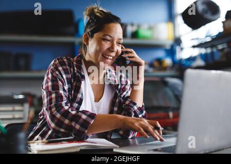 Smiling mixed race female car mechanic using smartphone and laptop in workshop Stock Photo