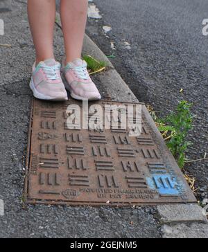 Man hole cover on the edge of a pavement. A blue dot of spray paint on the corner indicates it has been inspected. A child is standing at the edge Stock Photo