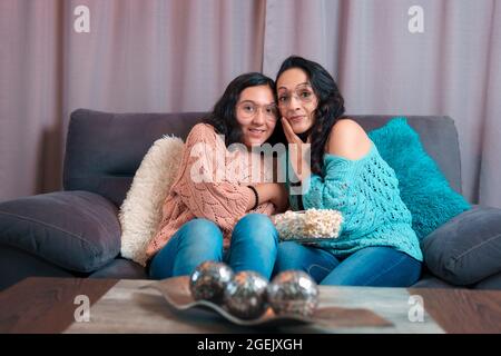 Horizontal view of a Mom and daughter watching a movie at home, they eat popcorn, they are both scared and make very different expressions Stock Photo