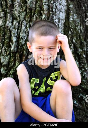 Beaming little boy smiles as he sits besides a tree outdoors.  He is leaning his head against his hand. Stock Photo