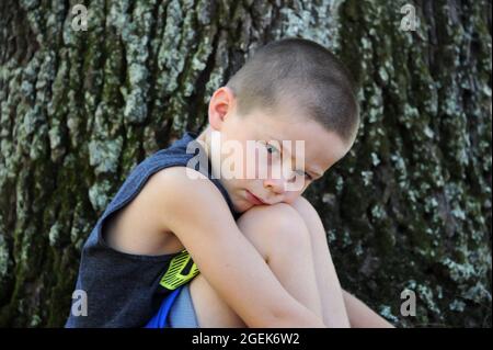 Little boy sits besides a tree outside.  He is sad because he doesn't have anyone to play with him.  He is hugging his knees and resting his head agai Stock Photo