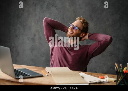 The smiling man in glasses sitting at the table with his hands behind his head in the studio Stock Photo