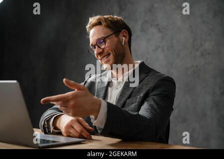 The smiling man in a jacket and glasses talking by video call on a laptop gesturing with his hands while sitting at the table in the office Stock Photo