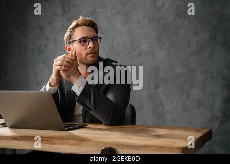 The confident man in suit and glasses looking to the side while sitting at the table with laptop in the studio Stock Photo