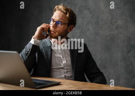 The pensive man in glasses talking on the phone and looking away while sitting at the table in the studio Stock Photo