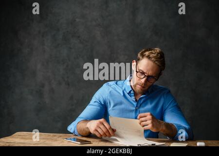 Frowning business man working with documents while sitting at the desk isolated over black background Stock Photo