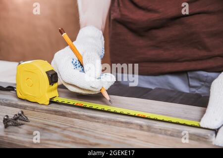 Exact measured. Close up of young male carpenter in gloves making measurements on the wooden plank by yellow measure tape. Stock Photo