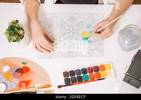 Art therapy or self expression for adults. Young woman is coloring page anti stress book, mental wellbeing concept. Workspace of artist - paints, penc Stock Photo