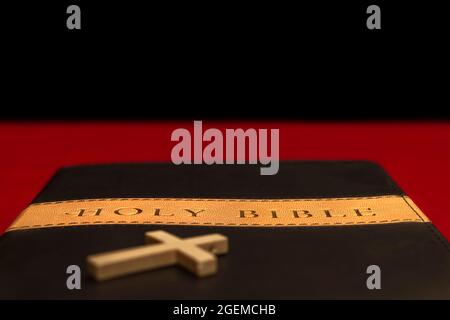 Holy bible on red table with a small crucifix in the foreground, with selective focus on the book's title. Stock Photo