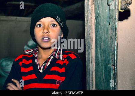 The Indian rural child is standing near the door of his house wearing a sweater and cap on a cold night. Stock Photo