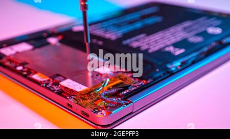 Unrecognizable man disassembles laptop with screwdriver in muted red and blue light Stock Photo