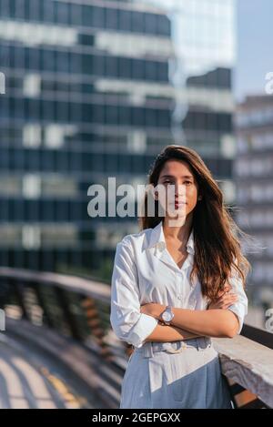 vertical portrait of a confident business woman looking at camera. She is crossing her arms and has offices buildings on the background Stock Photo