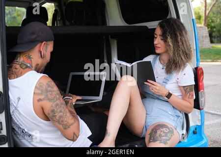 Young tattooed couple smiling, reading a book and working on a laptop sitting in the back of the van. Stock Photo