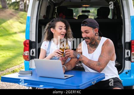 Young tattooed couple smiling, making a toast with beer bottles and using the laptop, sitting in a picnic table near by the van. Stock Photo