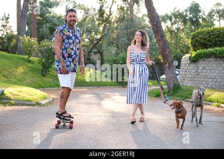 Young tattooed couple walking their dogs and riding a longboard. Stock Photo