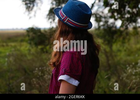 Defocus close up portrait of a serious tired young woman with brown hair wearing a hat outdoors. Green nature background. Back view. Out of focus. Stock Photo