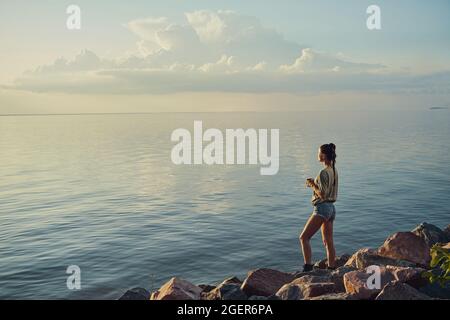 Adult standing on rocks with teacup and watching setting sun Stock Photo