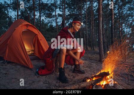 Female warming up near campfire while wearing blanket on shoulders Stock Photo