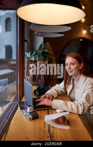 Smiling happy young female photographer sitting with laptop and camera at cafe. Concept of modern technology and photoartist. Side view portrait. Cauc Stock Photo