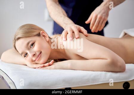 Charming Woman Lying On Belly Getting Spa Massage On Back By Male Masseur, Blonde Lady Of Caucasian Appearance Looking At Camera, Relaxed, Leisure Tim Stock Photo