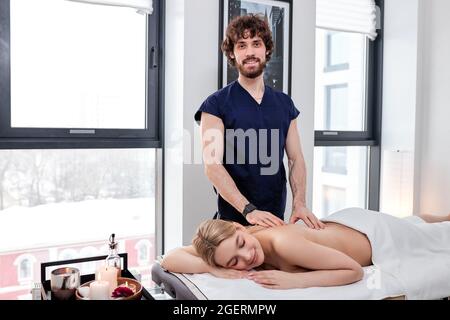 Portrait Of Bearded Nice Masseur In Uniform Carefully Doing Massage On Female Back, Blonde Caucasian Woman Lying On Belly Relaxing, Enjoying. Copy Spa Stock Photo
