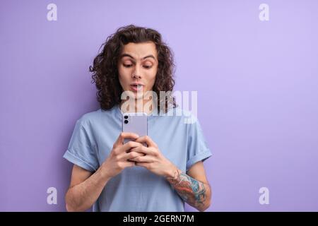 Young curly male emotionally reacts on message, surfing the Net, isolated portrait, purple background, People lifestyle, copy space concept Stock Photo