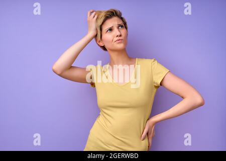 Confused short haired young woman touching head having dilemma difficult choice dressed in casual yellow t-shirt isolated over purple studio backgroun Stock Photo